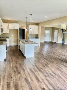 Open-concept kitchen and living area with wood-look flooring, white cabinets, granite countertops, an island with pendant lights, and neutral-colored walls. A doorway leads to another room near the entry door.