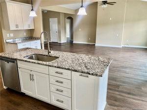 A modern kitchen with white cabinets, granite countertops, a stainless steel sink, and dishwasher, opening into a spacious living area with wood flooring, beige walls, and ceiling fan.