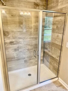 A glass shower enclosure with a metal frame features beige and brown rectangular tiles on the walls and a white shower base with a central drain. A chrome handle is attached to the door, and a window is visible outside.