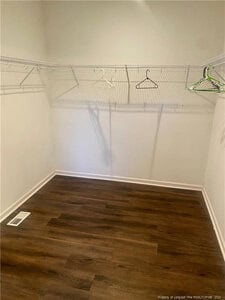 Empty walk-in closet with brown wood flooring, white walls, and white wire shelves along two walls. Several empty hangers are on the shelves. A floor vent is visible in the corner.
