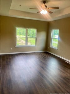 Empty room with tan walls, dark wood flooring, two large windows, one smaller window, and a ceiling fan with light. Natural light is coming through the windows, and the view outside shows greenery.