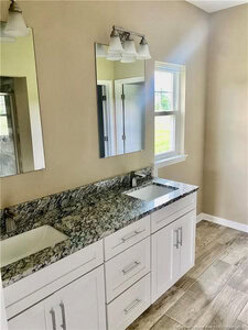 Double vanity with a granite countertop, two rectangular sinks, white cabinets, two mirrors, and light fixtures above. The bathroom has beige walls, a window, and wood-look tile flooring.