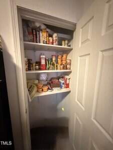 A small pantry with three shelves holding canned goods, boxed food, seasoning packets, instant noodles, and other pantry staples. The shelves are partially filled, and the pantry door is open.