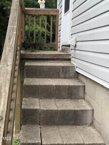 A set of six concrete steps with a wooden handrail leads up to a white door on the side of a house with gray siding. There is greenery in the background.