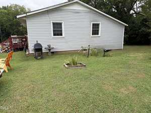 A backyard with green grass, a small concrete patio, a grill, a birdbath, chairs, and a raised garden bed in front of a white single-story house with two windows. Trees and a red deck are visible in the background.