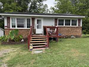 A one-story brick house at 206 1st Street, Oxford, features white siding, red shutters, and a front porch with red railings. Steps lead up to the porch with potted plants, lawn ornaments, and grass in the yard. Trees stand in the background.