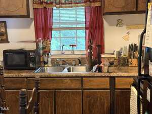 A kitchen sink with a double basin is set in a brown countertop. Above it is a window with red curtains and two small vases. To the left is a microwave; to the right are knives in a block and various utensils.