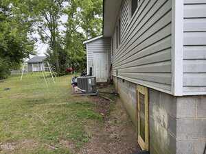 Side view of a house with gray siding and a small wooden crawlspace door; an HVAC unit, tools, and a storage shed are adjacent. The yard is grassy with a swing set and trees in the background.