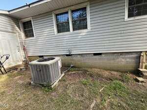 An outdoor air conditioning unit sits next to the exterior wall of a house with gray siding and multiple windows; garden tools lean against the wall near a white door. The ground is uneven with sparse grass.