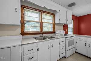 A kitchen with white cabinets, a double sink under a window with blinds, an electric stove, and beige countertops. The walls are partially red and beige, with natural light coming through the windows.