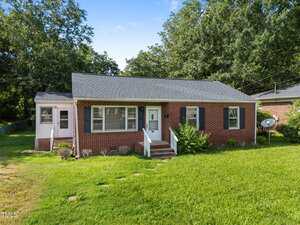 A single-story brick house with dark shutters, a small porch with steps, and a white front door. The lawn is grassy with stepping stones, and there are trees and another house nearby.