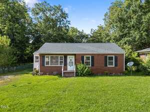 A single-story red brick house with white trim, black shutters, and a gray shingle roof sits on a grassy lawn. There are bushes near the front steps and trees in the background under a blue sky.