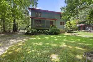 A two-story house with green vertical siding and red trim sits in a wooded area. The yard has green grass and mature trees, and a wooden shed is visible in the background to the right.
