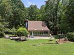 A modern house with a sloped roof sits in front of a wooded area at 4083 Graham Sherron Road. The neatly mowed lawn features landscaping, a stone patio, outdoor furniture, and a red umbrella in the foreground near Wake Forest.
