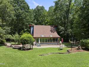 A modern house with a sloped roof sits in front of a wooded area at 4083 Graham Sherron Road. The neatly mowed lawn features landscaping, a stone patio, outdoor furniture, and a red umbrella in the foreground near Wake Forest.