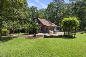 A two-story house with red trim sits in a grassy yard surrounded by tall trees. The backyard features a large patio, outdoor furniture, a fire pit, and a wooden pergola covered with greenery.