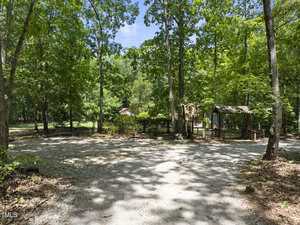 A gravel driveway surrounded by tall, green trees leads to a small wooden structure and a fenced area with a building partially visible in the background on a sunny day.