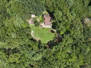 Aerial view of a house with a red roof surrounded by dense green trees and a large grassy yard, located in the center of a forested area. Paths and landscaping are visible near the house.