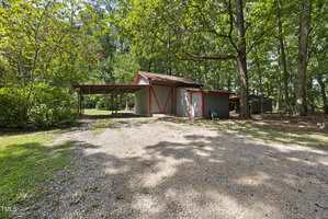 A gray and red barn-style shed sits in a wooded area with a gravel driveway. To the left is a covered carport. Shadows from tall trees cover the ground, and green grass borders the gravel driveway.