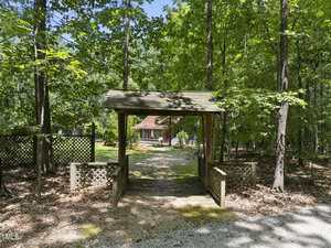 A wooden canopy structure stands over a gravel driveway leading to a house with a red roof, surrounded by green trees and shaded by forest. Lattice panels border the structure, and sunlight filters through the trees.