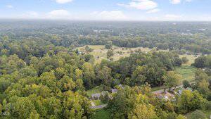 Aerial view of a rural area with dense green trees, scattered houses, and open grassy fields under a partly cloudy sky. A road runs through the foreground near several homes.
