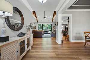 A spacious living area with wood floors, a sideboard with decor, ceiling beams, a large mirror, sectional sofas, and a view of a dining area and patio doors leading to a green outdoor space.
