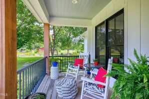 A covered porch with two white rocking chairs, a small round table, potted plants, and red cushions. The porch overlooks a grassy yard with trees and a red outbuilding in the background.