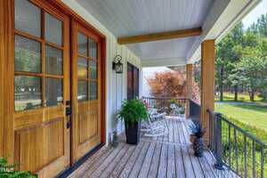 A covered wooden porch with two white rocking chairs, a potted fern, and a pineapple decoration. The porch has wooden doors with glass panes and overlooks a green yard with trees and shrubs.