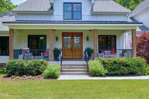 A modern farmhouse with white siding, a covered front porch, wooden double doors, steps leading up, black railings, green shrubs, and a well-maintained lawn. There are chairs and potted plants on the porch.