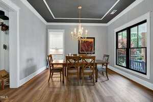 A dining room with light gray walls, a dark gray ceiling, and wood flooring. A wooden dining table with six chairs is centered under a chandelier. Large windows and a wall painting are visible. A kitchen is seen through a doorway.