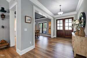 A view of a home's entryway with wood flooring, double glass front doors, a light fixture, a sideboard with decorative vases and plants, and a dining area visible through an open doorway.