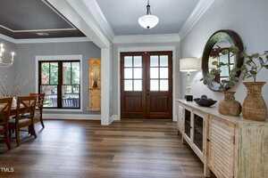 A bright entryway with wooden double doors, a sideboard with decor items, a round mirror, and a table lamp. To the left, a dining area with a wooden table, chairs, and large windows is visible.
