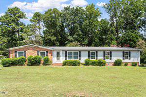 Charming single-story ranch-style house at 65 Pine View Road, Henderson, with a brick and siding exterior, six front windows, a white front door, and small shrubs along the facade. Set on a grassy lawn with trees in the background.