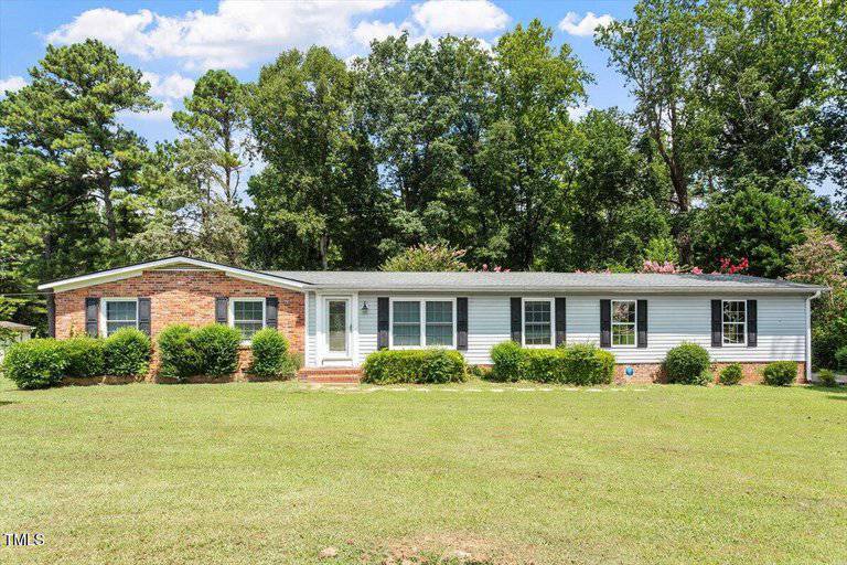 Charming single-story ranch-style house at 65 Pine View Road, Henderson, with a brick and siding exterior, six front windows, a white front door, and small shrubs along the facade. Set on a grassy lawn with trees in the background.