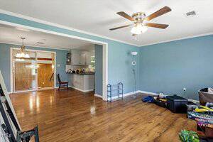 A living room with light blue walls, hardwood floors, a ceiling fan with light, and scattered items on the floor. The space opens to a kitchen and dining area with sliding glass doors in the background.