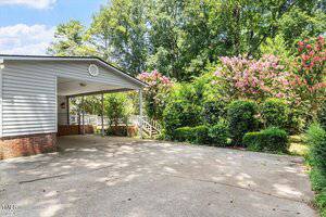 A concrete driveway leads to a carport attached to a house with white siding and brick trim. Trees, shrubs, and flowering bushes line the driveway and surround the area.