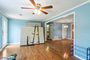 A room with light blue walls, wooden floor, and a ceiling fan with lights. A mattress and bed frame are propped against the wall. A metal shoe rack is in the corner, and a door with a glass window is on the left.