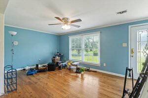 A mostly empty room with blue walls, wood floors, a ceiling fan with light, two large windows with blinds, and scattered items on the floor including bags, boxes, and small furniture pieces.