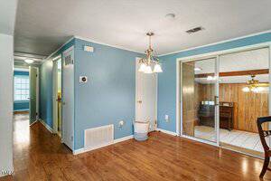 A dining area with light blue walls, wooden floors, and a brass chandelier. There is a hallway on the left, a closed white door in the center, and large sliding glass doors opening to a wood-paneled room.