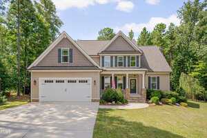 Located at 3800 Watermark Drive in Franklinton, this two-story suburban house features beige siding, stone accents, a front porch, and a two-car garage, nestled among green trees and landscaped shrubbery with an American flag by the entrance.