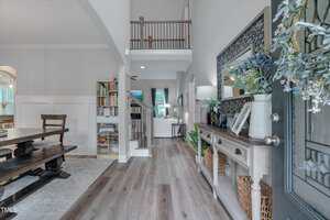 A view of a home entryway with light wood floors, a console table with decor and baskets, a staircase leading upstairs, bookshelves, a dining area to the left, and a living room visible in the background.
