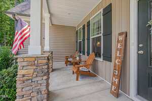 A front porch with stone columns, two wooden chairs, a small table, a large "Welcome" sign, and an American flag. The house has tan siding, black shutters, and greenery visible in the background.