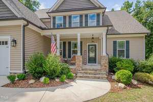 A two-story house with tan siding, stone porch columns, and white trim. The front porch has a welcome sign, two chairs, and an American flag. The landscaped yard includes bushes and a curved walkway.