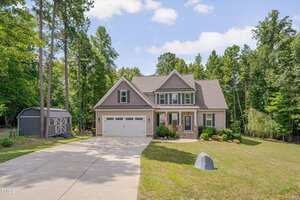 A two-story suburban house with gray siding, a gable roof, and an attached double garage is shown with a neatly landscaped front yard, a concrete driveway, and a gray storage shed on the left. Tall trees surround the property.