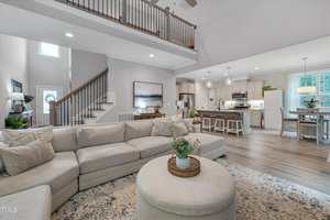 Spacious open-concept living area with a large beige sectional sofa, round ottoman, and kitchen with white cabinets and an island. Wooden stairs and a balcony railing are visible in the background. Natural light fills the room.