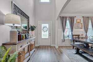A bright entryway with a white front door, a sideboard table holding a lamp, baskets, and framed photos. To the right, a dining area with a wooden table and chairs is visible, featuring blue curtains and wall decor.