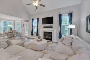 A spacious living room with a beige sectional sofa, round ottoman, and patterned rug. There is a fireplace with a TV mounted above it, large windows with blue curtains, and a ceiling fan. A dining area is visible in the background.
