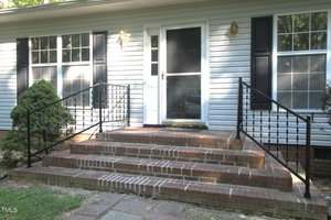 A front entrance with brick steps leading to a black storm door and white siding. There are black metal railings on both sides, two windows with black shutters, and two outdoor wall lanterns on either side of the door.