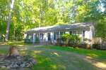 Charming single-story house at 622 Enon Road, Oxford, with light-colored siding, black shutters, and a dark roof. Enjoy a front porch with railings, bushes along the front, and a tree stump in the grassy yard surrounded by trees.