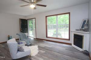 A living room with gray wood flooring, a ceiling fan, large window, and open door. The space has a patterned chair, a small white rug, a wrapped blanket, and a corner fireplace with a framed photo and decor on top.